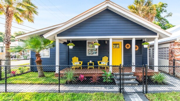 A house with a dark blue exterior, a yellow door, and a porch with two yellow Adirondack chairs and a small table. - Historic Cottage Walk to Downtown St Augustine Free Parking Sleeps 4 (St. Augustine)