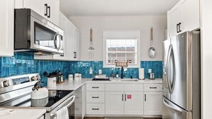 A modern kitchen with white cabinets, a stainless steel refrigerator, a microwave, and a stove. There is a window with blinds and a blue tiled backsplash. - Historic Cottage Walk to Downtown St Augustine Free Parking Sleeps 4 (St. Augustine)