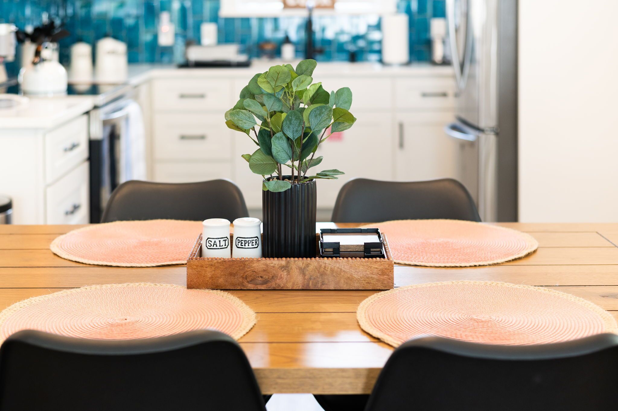 Una mesa de madera para comer con dos individuales, un florero negro con follaje verde y saleros y pimenteros.
