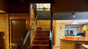 Una escalera de madera que conduce a un segundo piso, una zona de cocina con barra y un comedor con una mesa y sillas de madera.