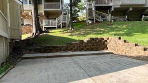Un carrer d'accés de formigó que condueix a una entrada tancada per un mur de maó i una zona verda amb arbres.