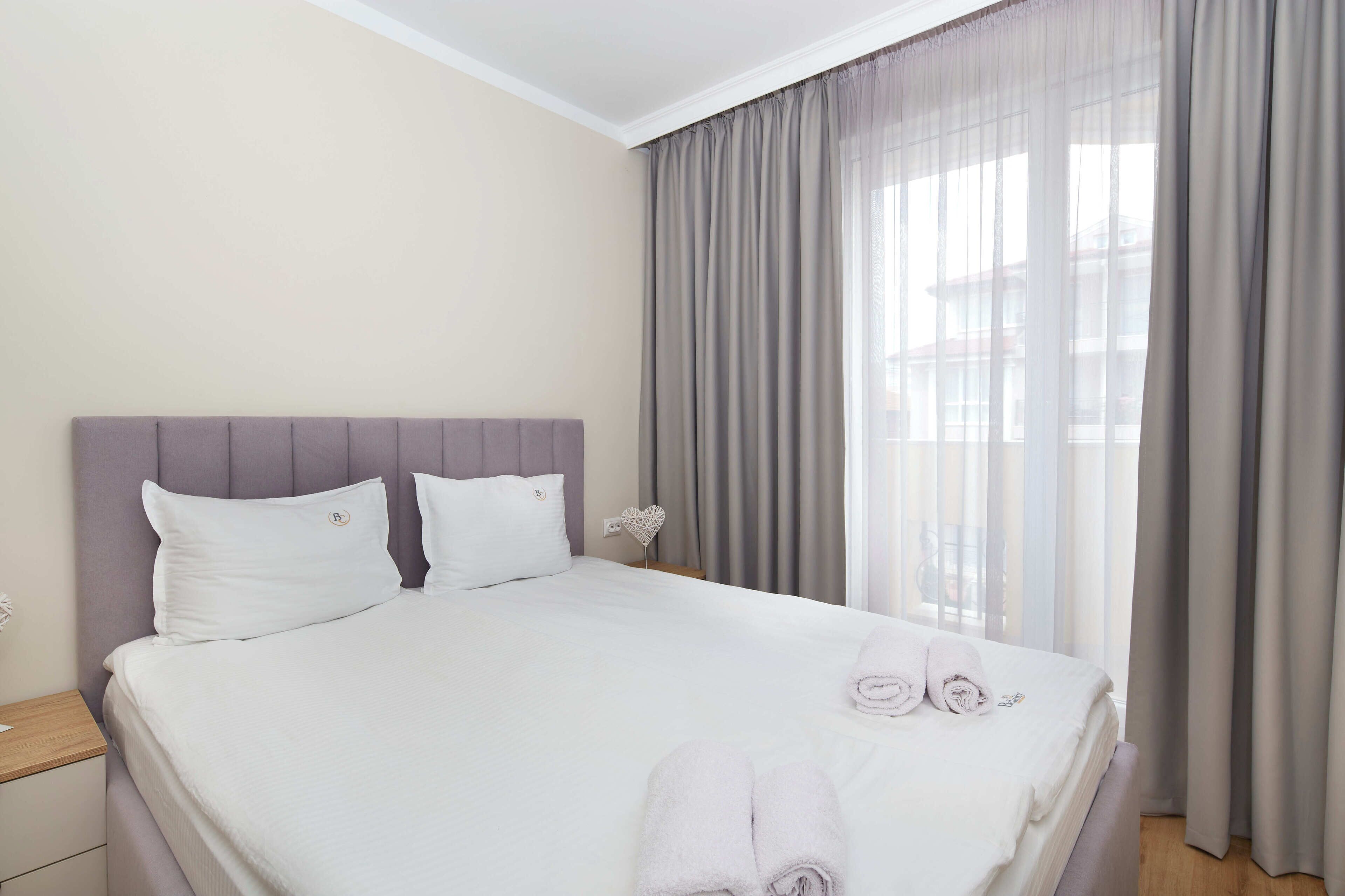 Un lit bien fait, avec des draps blancs et deux oreillers, une table de chevet avec une lampe, et une vue sur l’extérieur à travers des rideaux légers.