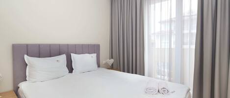 Un lit bien fait, avec des draps blancs et deux oreillers, une table de chevet avec une lampe, et une vue sur l’extérieur à travers des rideaux légers.