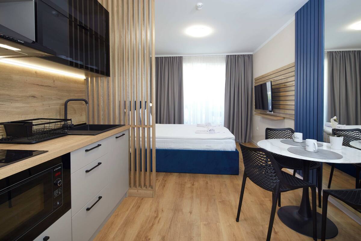 A compact hotel room with a kitchenette, a bed, a dining area, and a TV mounted on the wall.