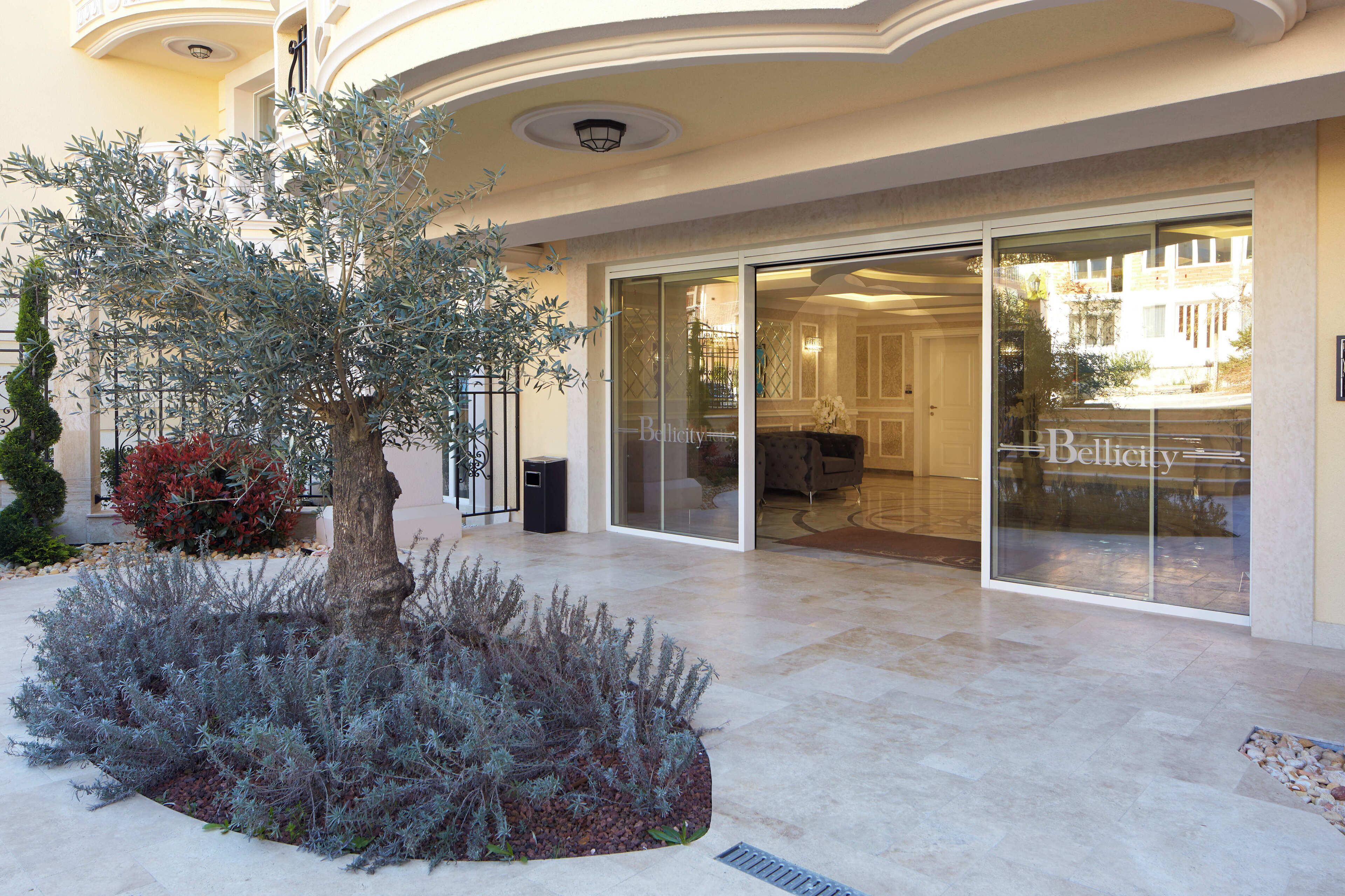 A hotel entrance with a large glass door, a well-maintained garden with a tree and shrubs, and a clean, tiled floor.