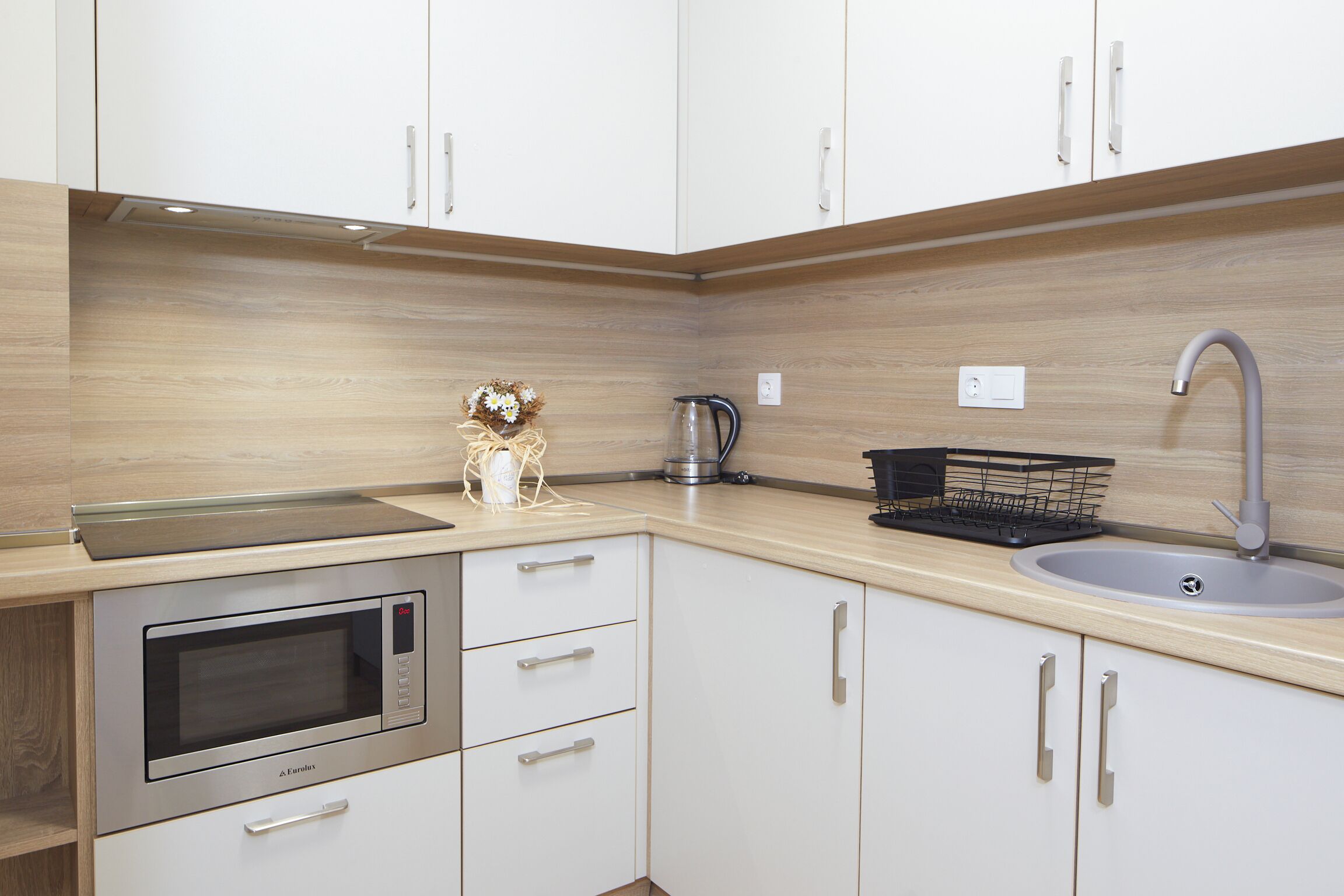 A modern kitchen with white cabinets, a built-in microwave, a kettle, and a sink.