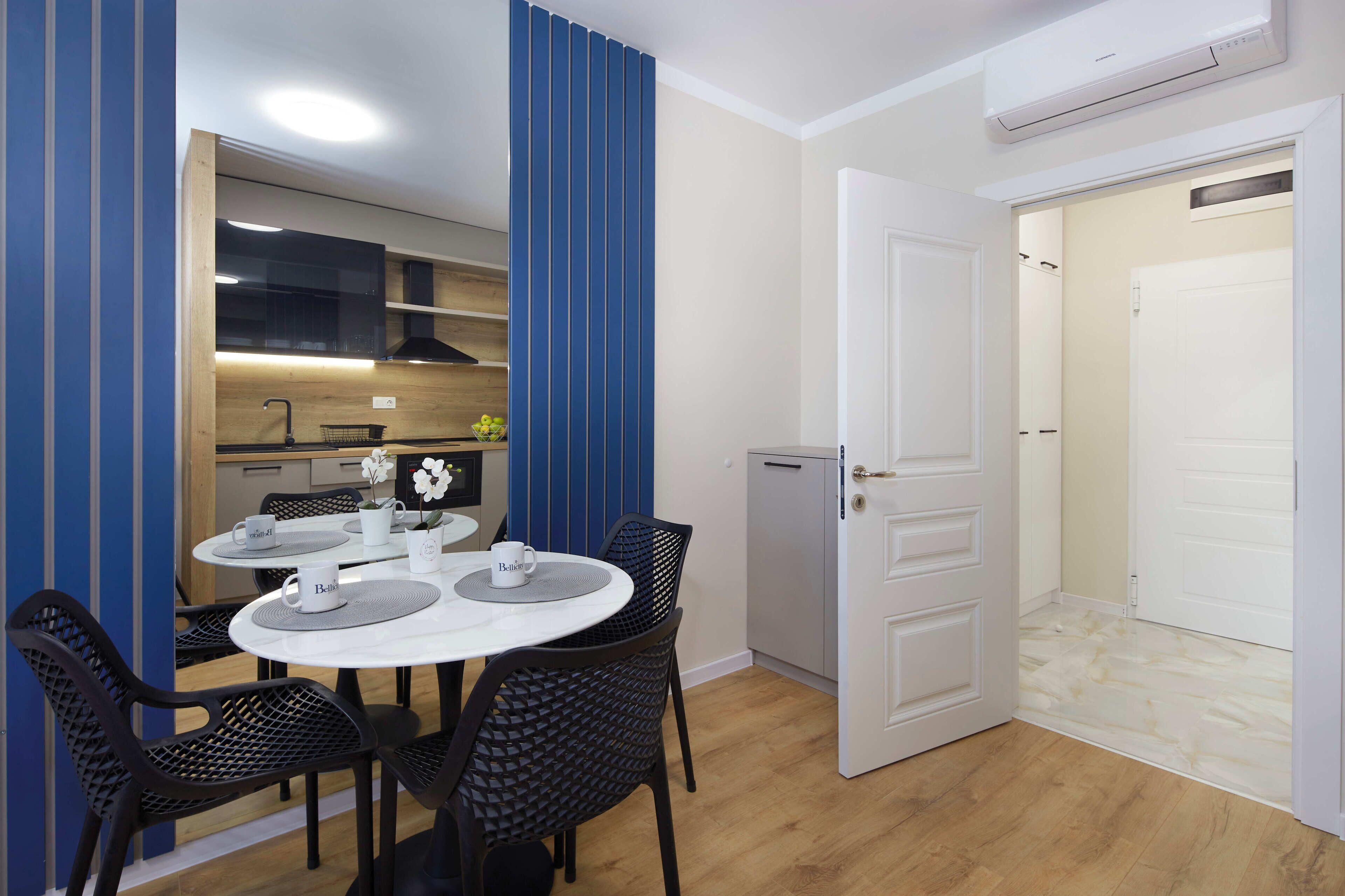 Una cucina compatta con elettrodomestici moderni, un'area pranzo con tavolo e sedie rotonde, e una porta bianca che conduce al bagno.