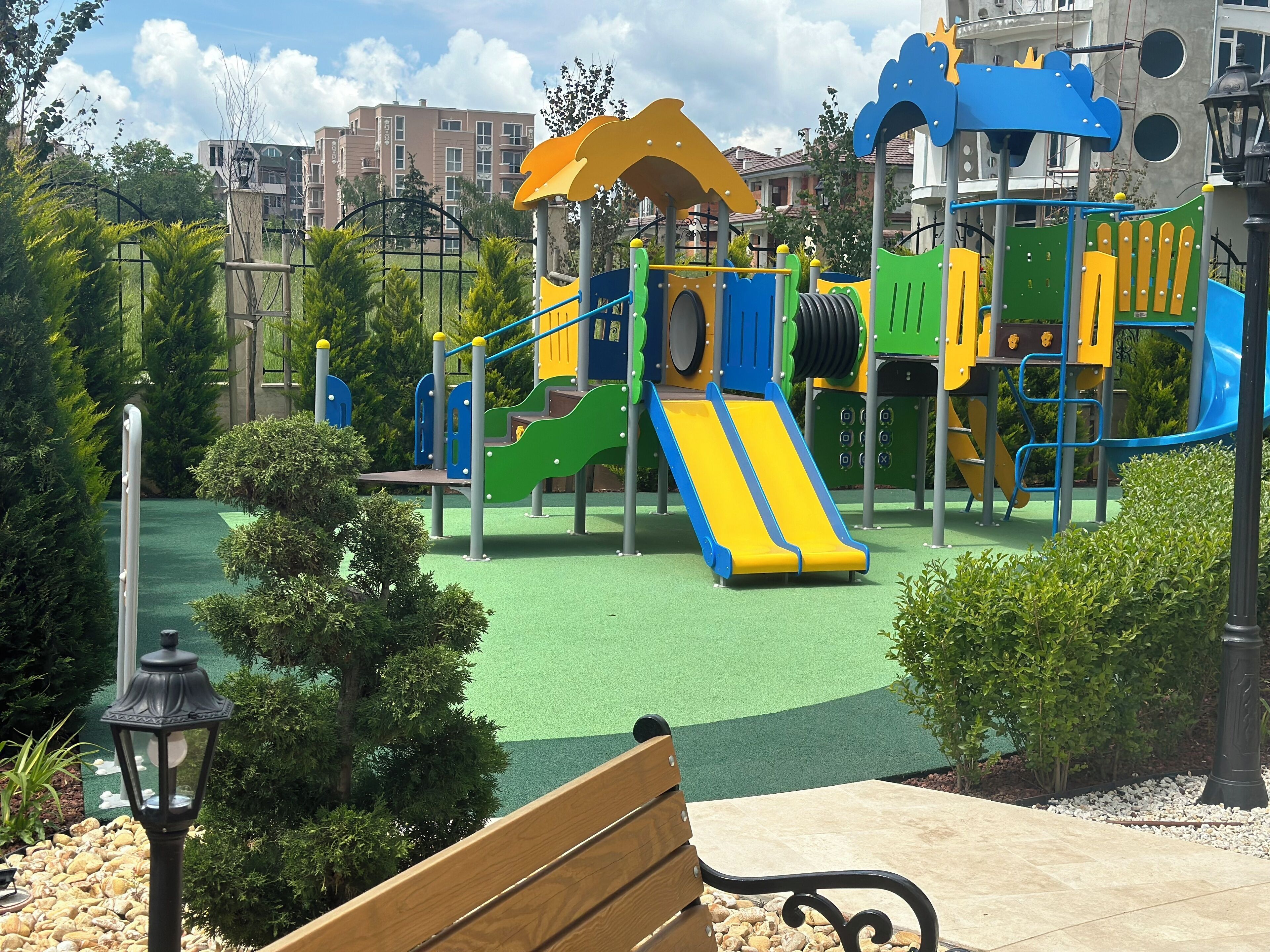 A playground with a slide and climbing structures, surrounded by greenery and a paved area with benches.
