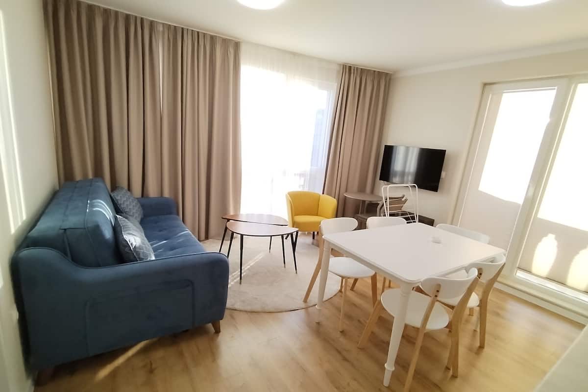 A living room with a blue sofa, a yellow armchair, a small round table, a white dining table with chairs, a flat-screen TV mounted on the wall, and large windows with curtains.