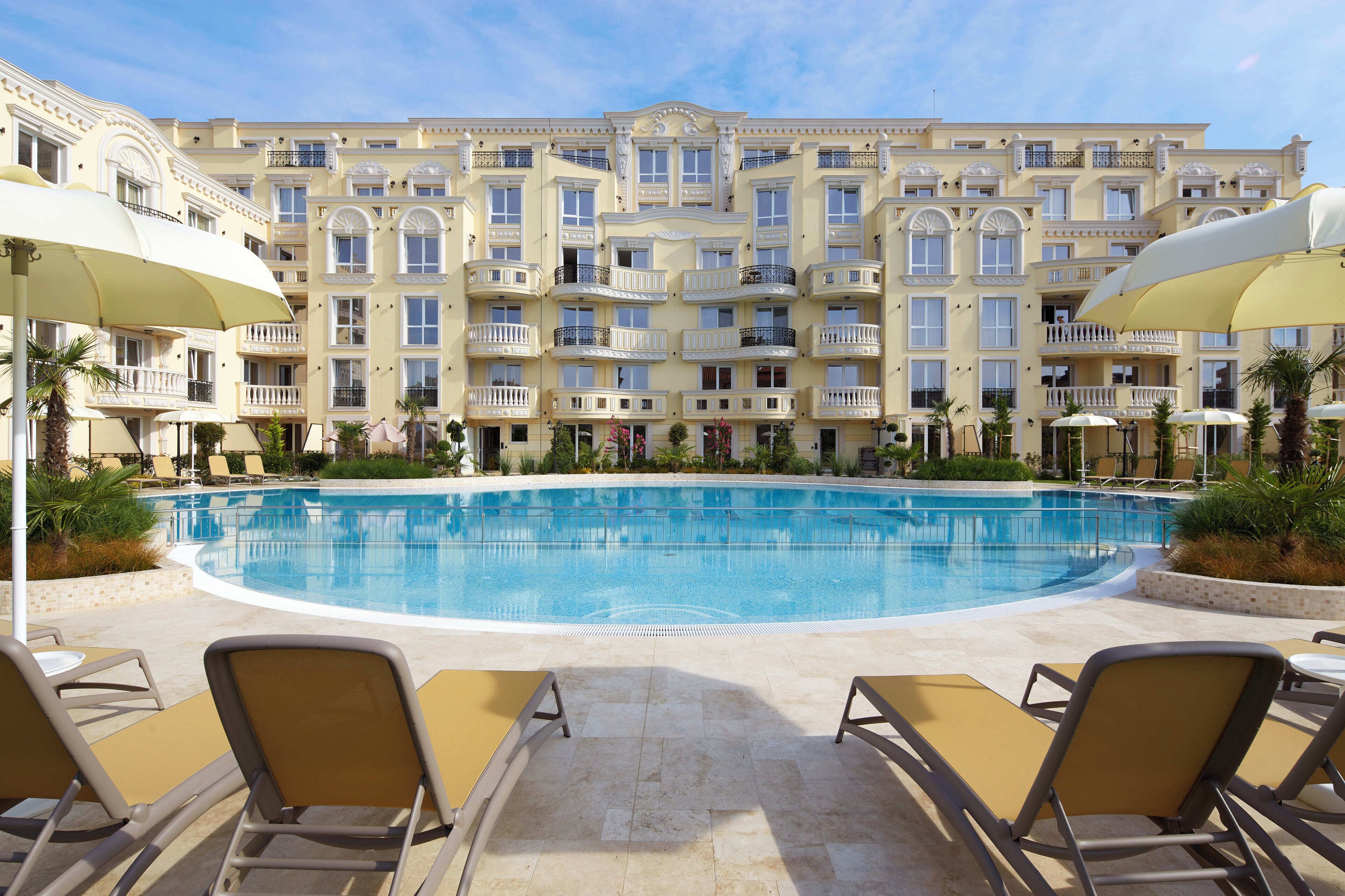 A large pool surrounded by sun loungers and umbrellas, with a multi-story building in the background.