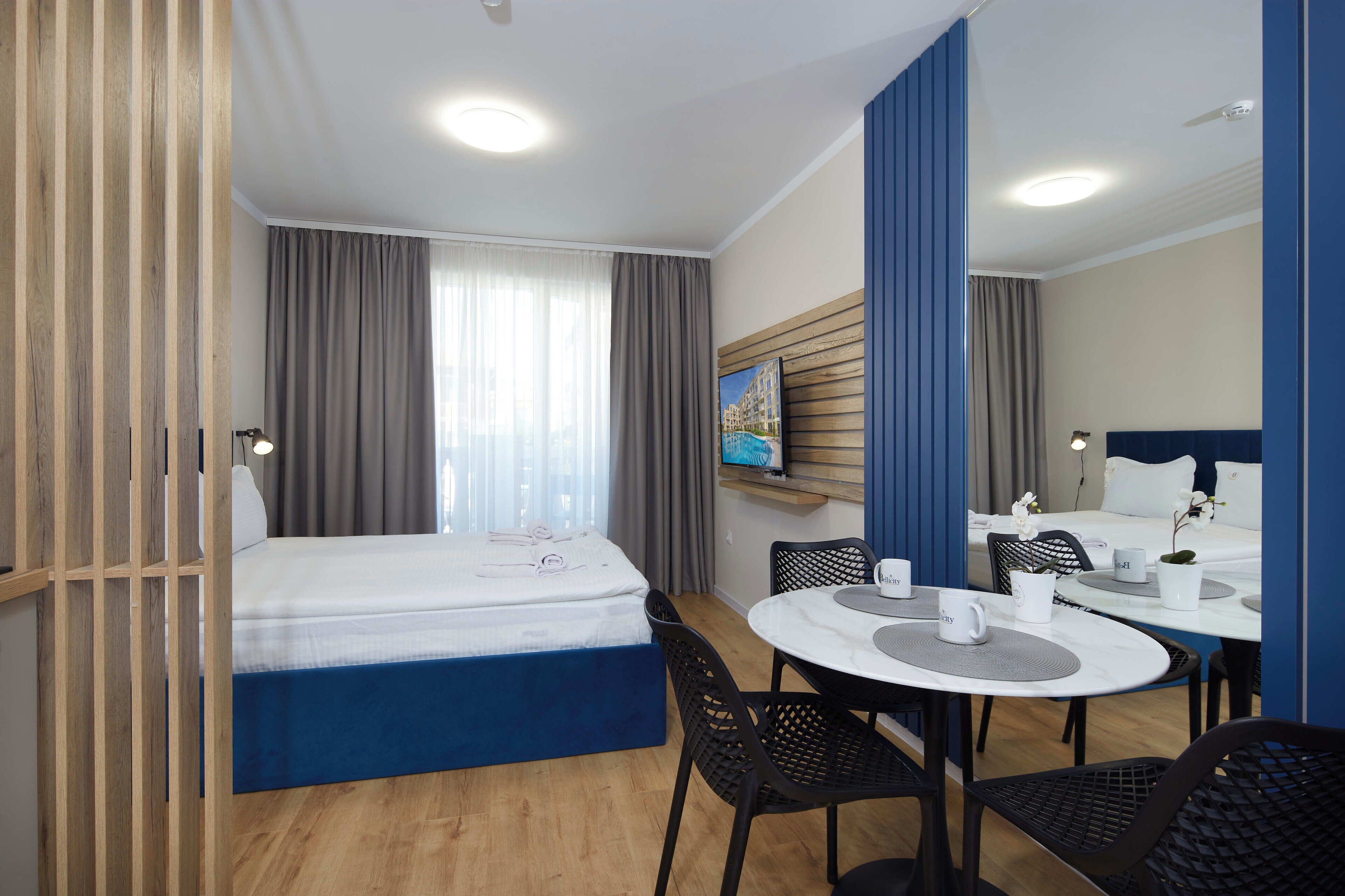 Une chambre d’hôtel comprenant un lit, une table à manger avec des chaises, un petit coin cuisine et une grande fenêtre avec des rideaux.