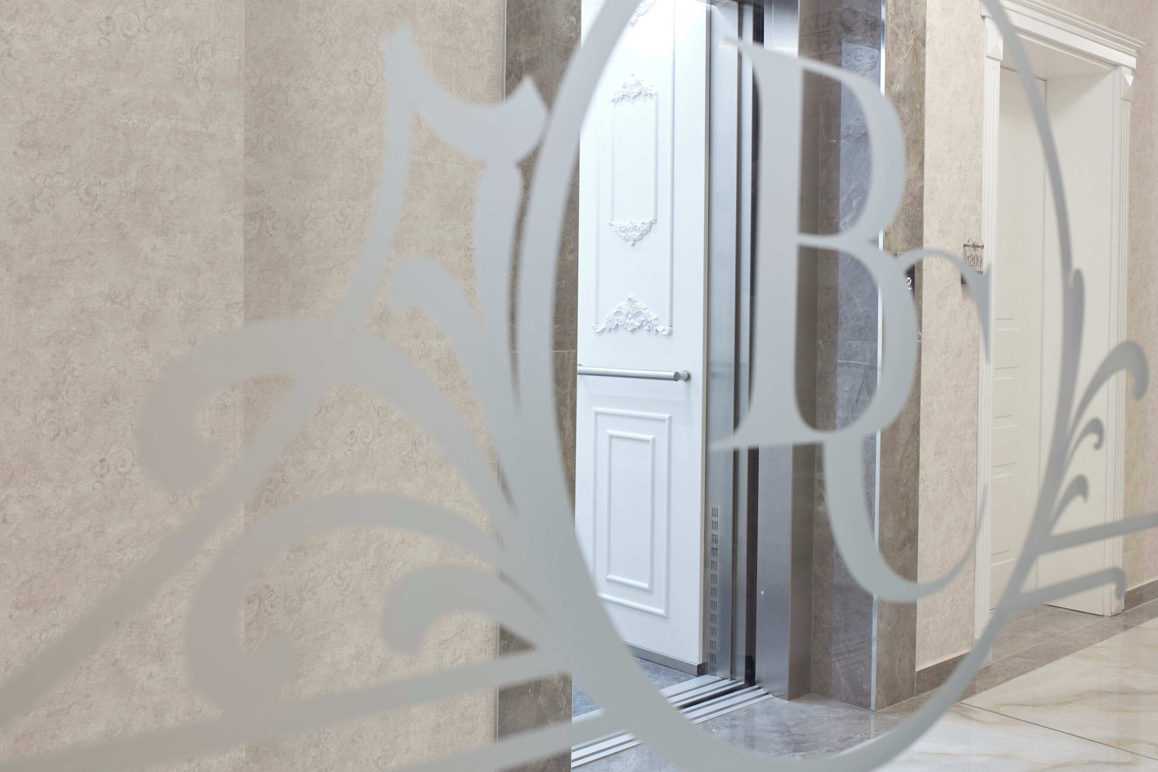 Un pasillo con suelo de mármol, una puerta blanca con manija de metal y un letrero decorativo de metal con un monograma.