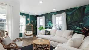 Un salon moderne avec un canapé blanc, des fauteuils en osier, une table basse ronde en osier et un mur décoré d’une fresque représentant des feuilles vertes.
