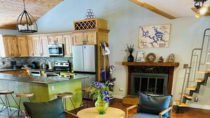 Uma sala de estar ampla com piso de madeira, uma mesa de jantar com cadeiras, uma lareira e uma cozinha ao fundo.