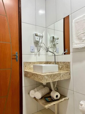 A bathroom with a white sink, two hair dryers mounted on the wall, a towel rack with towels, and a shelf with a towel and a roll of toilet paper.