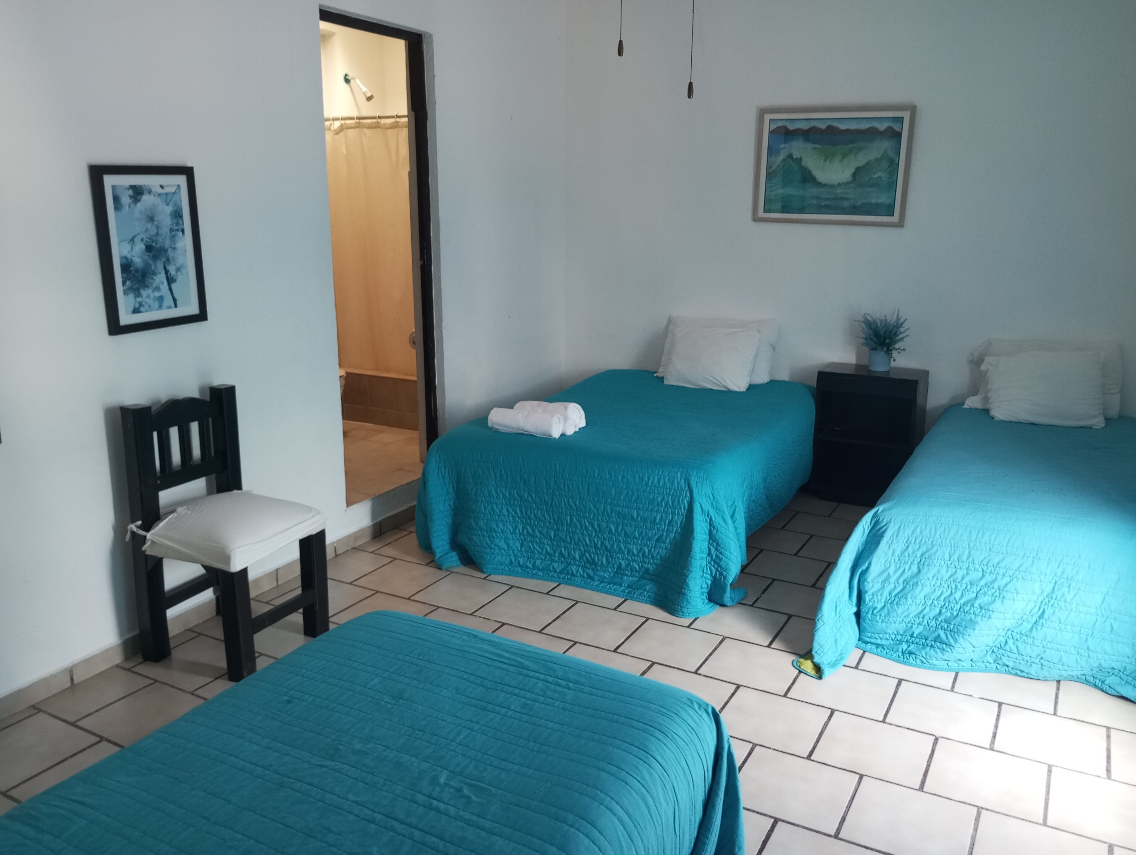 A room with two beds, a chair, a nightstand, a painting, and a door leading to a bathroom.