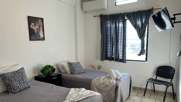 A hotel room with two beds, a chair, a desk, a plant, and a painting on the wall. - Casa Los Angeles  (Colima)