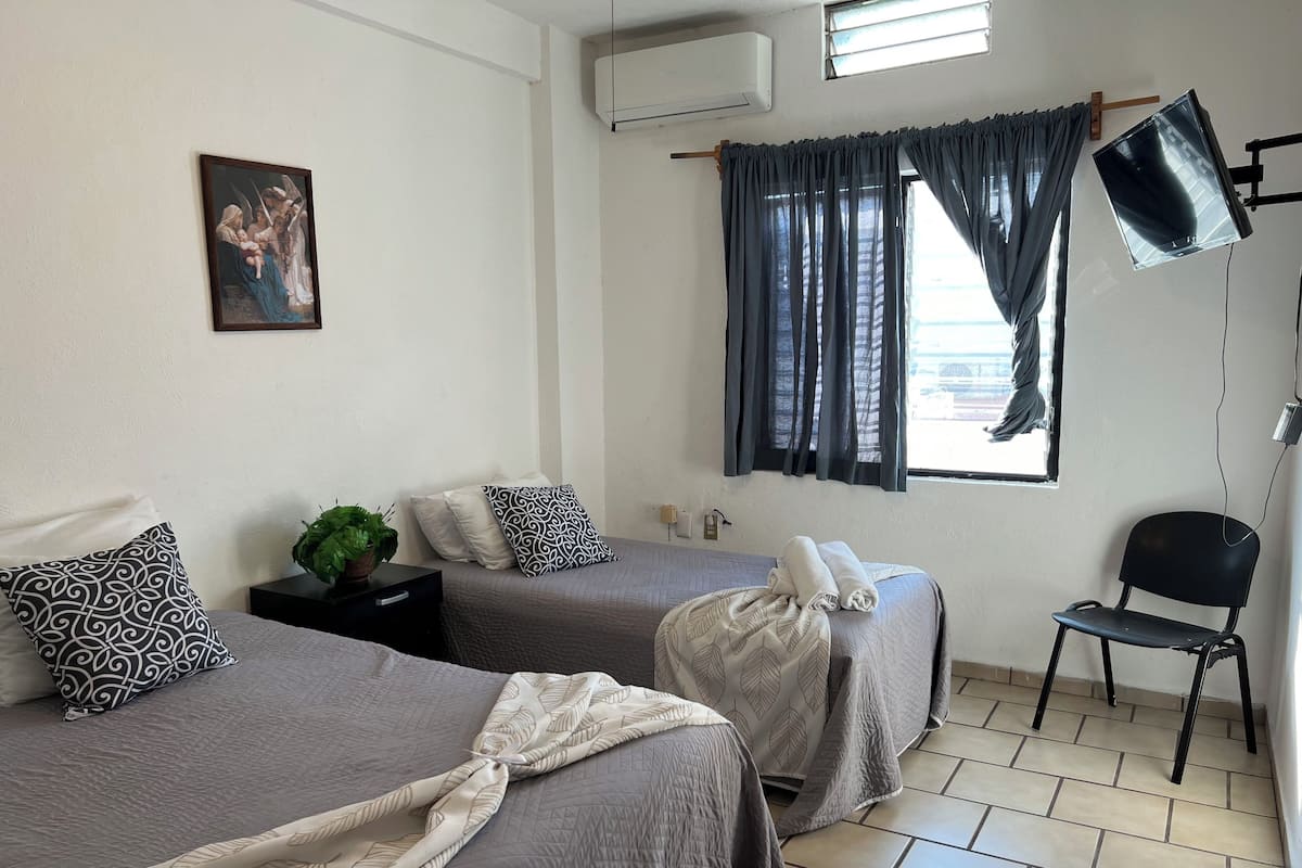 A hotel room with two beds, a chair, a desk, a plant, and a painting on the wall.