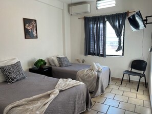 Ein Hotelzimmer mit zwei Betten, einem Sessel, einem Schreibtisch, einer Pflanze und einem Bild an der Wand.