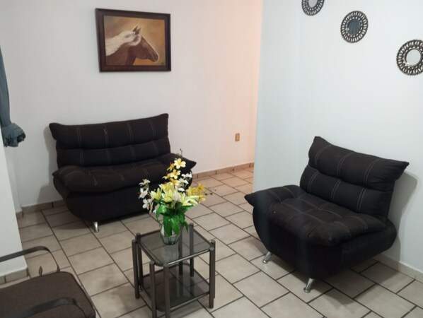 Una sala de estar con un sofá marrón oscuro, una mesa de centro de cristal con un jarrón de flores y un cuadro enmarcado en la pared.