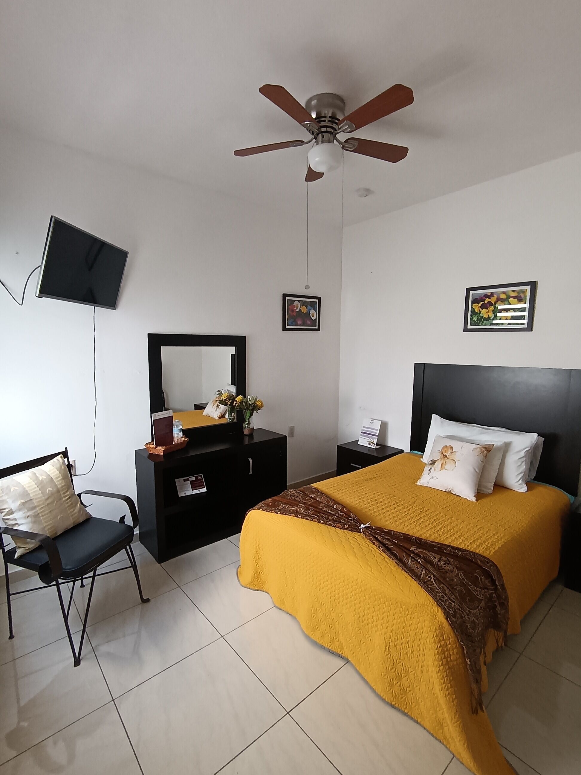 Une chambre à coucher comprenant un lit, un bureau, une chaise, un ventilateur de plafond et un téléviseur fixé au mur.