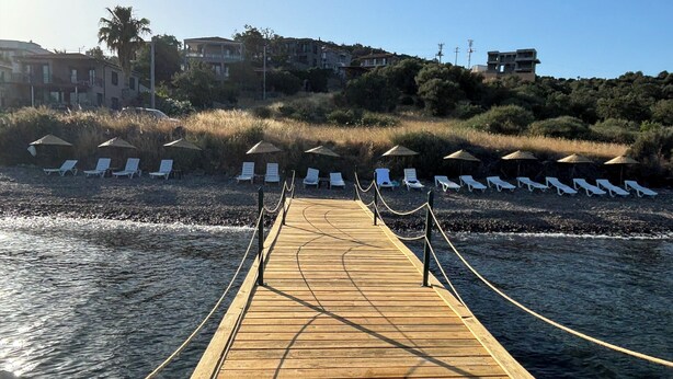 Un muelle de madera que lleva a sillones de playa con sombrillas, con vistas a un mar tranquilo y una colina con edificios.
