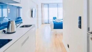 A modern, well-lit hotel room with a kitchenette, a bed with blue bedding, and a view of the room through a window. - orbi city 777 (Batumi)