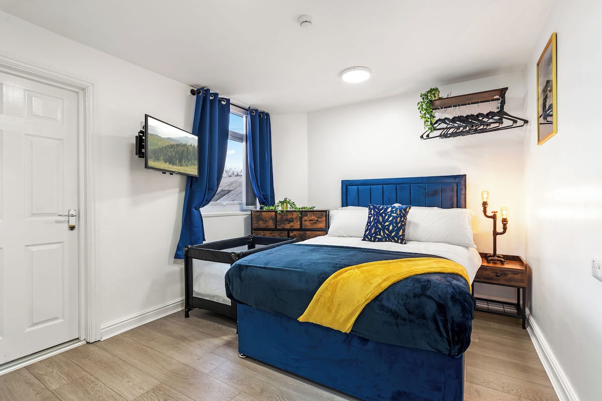 A bedroom with a bed, a bedside table, a wall-mounted TV, a window with curtains, and a wall-mounted plant rack.