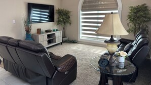 A living room with a leather sofa, a glass coffee table, a TV mounted on the wall, and a ceiling fan.