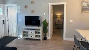 Ein Wohnzimmer mit Fernseher auf einer weißen Unterhaltungsecke, ein Essbereich mit Tisch und Stühlen sowie ein Flur, der in ein weiteres Zimmer führt.