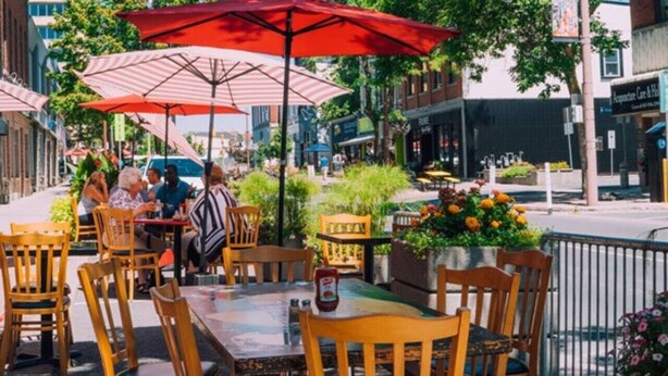 Een terras met tafels en stoelen onder gestreepte parasols, omringd door groen en gebouwen.