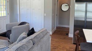 A modern living room with a grey sofa, a dining table, and a kitchen area with white cabinets and a stainless steel refrigerator. - Brand New Stylish Black Cottage (South Kingstown)