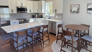 A modern kitchen with white cabinets, stainless steel appliances, a central island with bar stools, and a round dining table. - Brand New Stylish Black Cottage (South Kingstown)