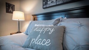 A neatly made bed with a pillow that reads "happy place" and a lamp on a bedside table.