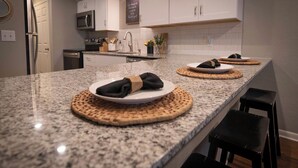 A kitchen with granite countertops, white cabinets, and a stainless steel refrigerator. There are two woven placemats on the counter, and a black stool is visible.