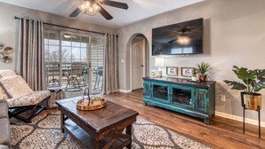 A living room with a ceiling fan, a TV mounted on the wall, a teal cabinet, a dining table, and a large window with curtains.