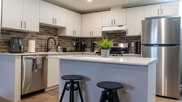 A modern kitchen with white cabinets, stainless steel appliances, a brick backsplash, and two bar stools. - Cozy 2BDR Lots Of Amenities (Cornwall)