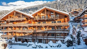A multi-story wooden chalet with balconies, surrounded by snow-covered trees and mountains.