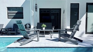 A modern outdoor pool area with lounge chairs and a table, surrounded by a white building with a glass door. - The Miami Grove Oasis (Miami)
