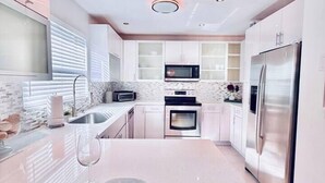 A modern kitchen with white cabinetry, stainless steel appliances, and a large island with a wine glass and plates. - The Miami Grove Oasis (Miami)