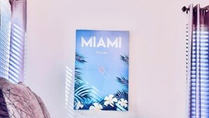 A hotel room with a bed, a pillow, a wall-mounted poster of Miami, Florida, and a window with blinds. - The Miami Grove Oasis (Miami)