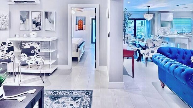 A modern living room with a blue sofa, a coffee table, and a patterned rug. There is a dining area with a chandelier and a kitchen visible in the background. - The Miami Grove Oasis (Miami)