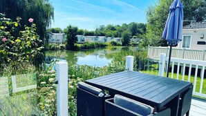 A deck with a table and chairs overlooking a lake, surrounded by greenery and a white fence.