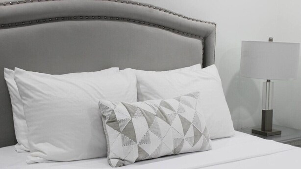 A bed with white bedding and two pillows, one plain and one with a geometric pattern.
