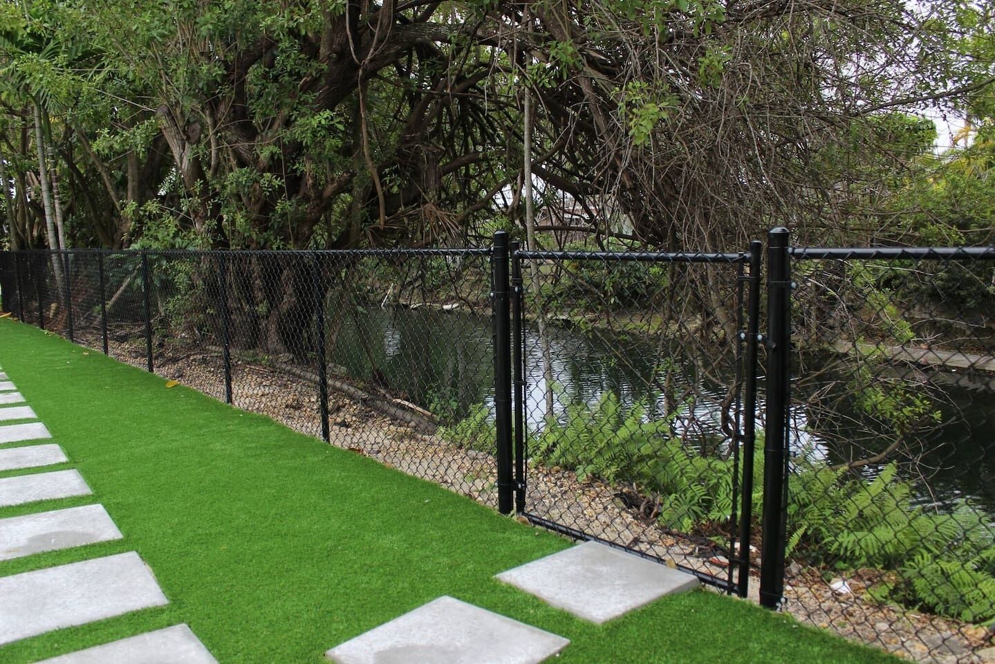 A fenced pathway with artificial grass leading to a body of water, surrounded by dense foliage.