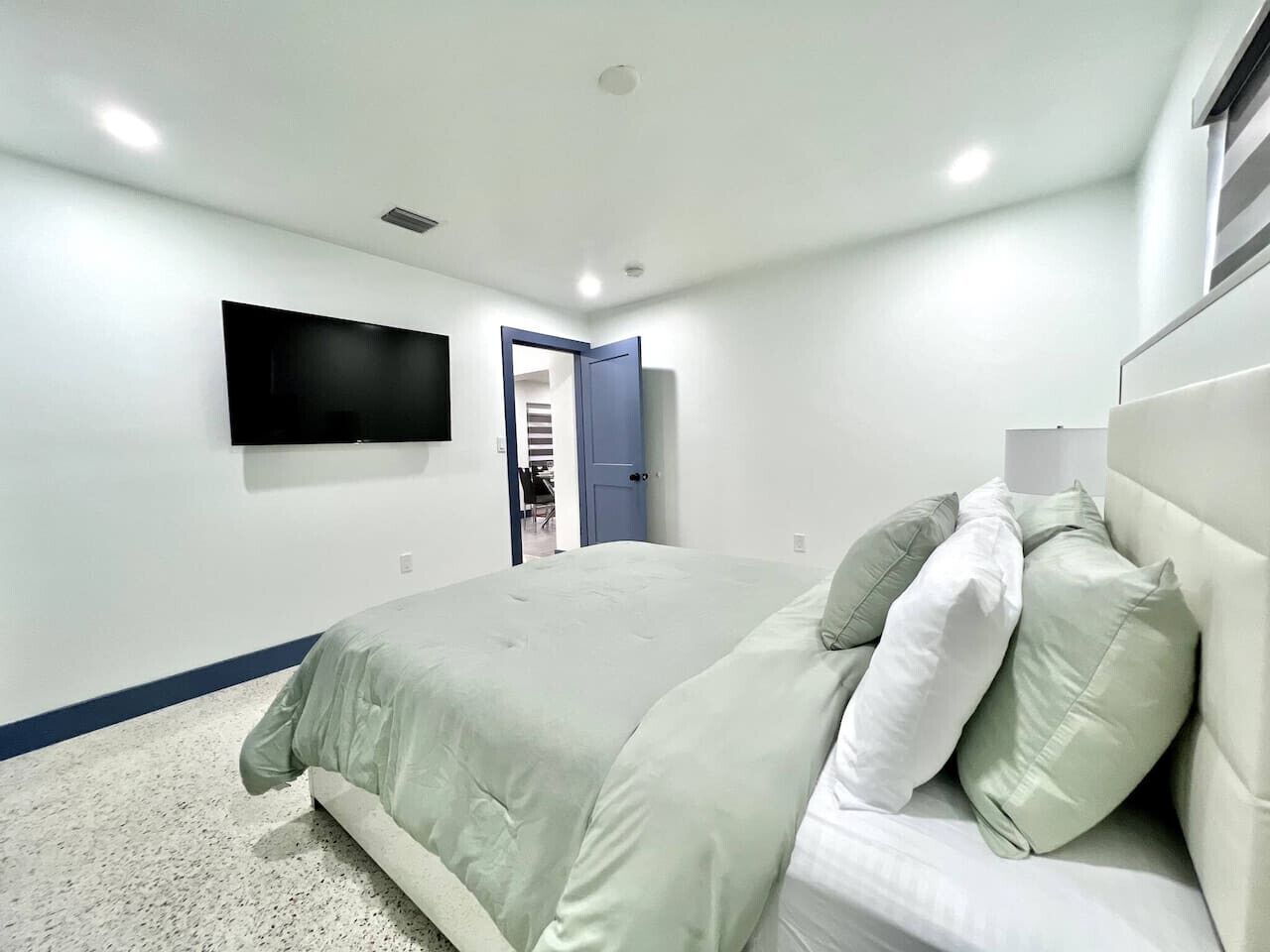 A bedroom with a bed, a flat-screen TV mounted on the wall, and a door leading to another room.