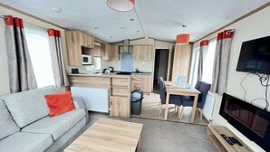 A compact living space with a sofa, a dining table, a TV, and a kitchenette. - Costwold Lodge, dog friendly with 3 bedrooms (Hoburne Holiday Park)