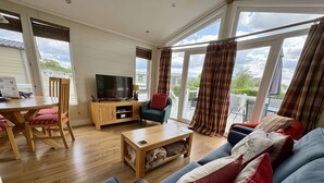 A spacious living room with a wooden dining table, a sofa, a television, and large windows with curtains.