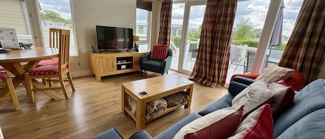 A spacious living room with a wooden dining table, a sofa, a television, and large windows with curtains.