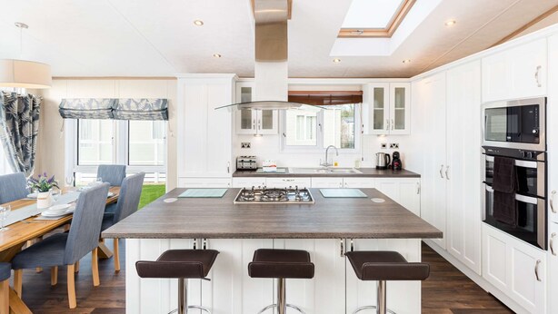 A modern kitchen with a central island, bar stools, a dining area with a table and chairs, and a view of the outdoors through large windows.